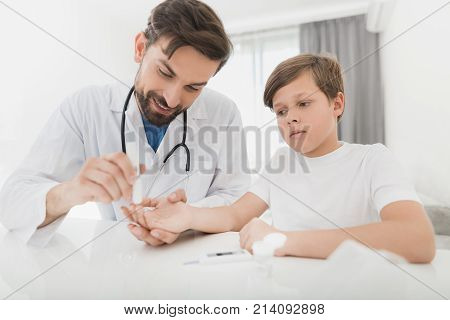 The doctor takes a blood sample from the boy to check it for sugar. The boy patiently endures the procedure. The doctor holds a scarifier in his hands. They are in a modern clinic.