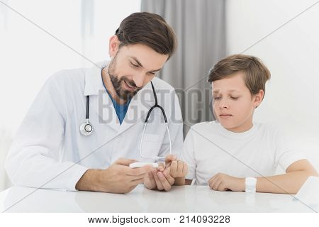The doctor takes the blood from the boy with a finger using a scarifier. The boy staunchly endures the procedure. The doctor will check the boy's blood for sugar level.
