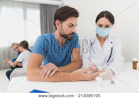 A nurse takes a blood sample from a man with a scarifier. A nurse is sitting in a mask and gloves at the table. They are in a man's house. Behind sit an old man and a boy.