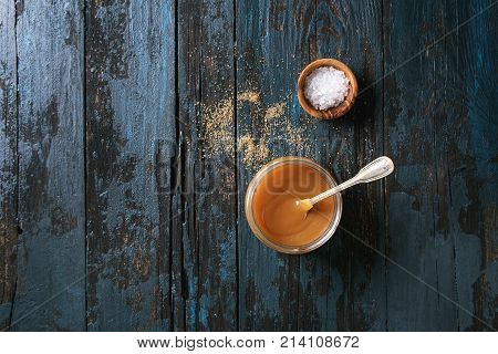 Glass jar of homemade salted caramel sauce with spoon, brown sugar and bowl of salt. Over old dark blue wooden background. Top view with space