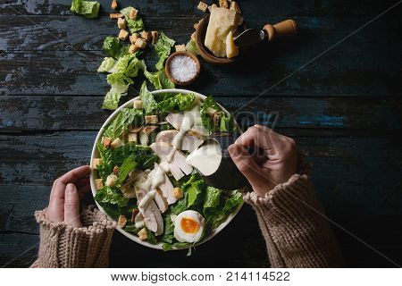 Female hands powring dressing to Classic Caesar salad with chicken breast in white ceramic plate. Served with ingredients above over old dark blue wooden background. Flat lay. Rustic style