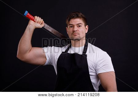 A sturdy man holds a machete by the shoulder in a black apron on a black background