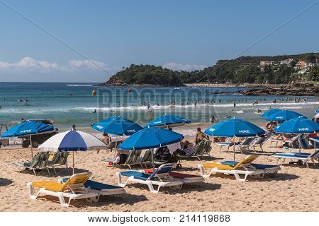 Sydney Australia - March 26 2017: Section of Manly Beach with sand and Tasman Sea. Series of beach chairs for hire with blue umbrellas. People on the sand and in the water. Small Shelly beach in distance.