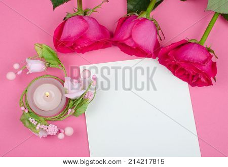 Beautiful pink roses and a burning candle on a blank white greeting card against a bright pink background (flat lay top view)