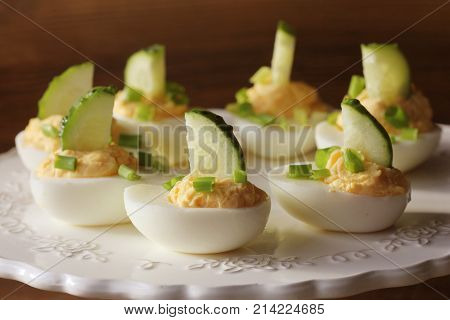 Spicy deviled eggs garnished with cucumber and leek on white plate. Wooden rustic background .