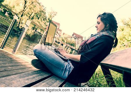 Young woman using laptop at a riverbank. Milleanial working in a park.