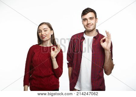 Isolated shot of cute cheerful young couple man and woman advertising some product making ok gesture as sign of good quality posing at studio wall. People goods advertisment and family concept