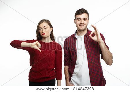 Displeased young woman in sweater showing thumbs down frowning having grumpy expression while posing in studio next to her happy husband who is expressing opposite emotions making ok sign