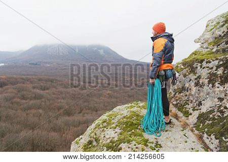 fully equipped The hipster-climber is standing in a down jacket and sneakers on a high rock with a coiled cove in a hand and a bag for chalk and magnesia on a belt with carbines. The concept of extreme rest in the mountains