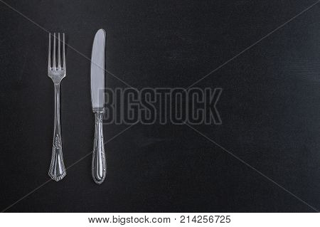 Fork and knife on black chalkboard background top view template for menu. Vintage silver tableware with copy space.