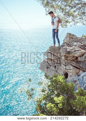Traveler standing near steep on the rocks near the sea under a tree and looking down. Summer Travel Vacation. Side view of handsome young caucasian tourist man in casual clothes outdoors on the nature.
