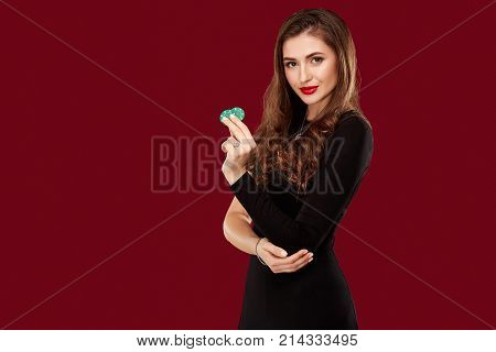Pretty long hair woman in black dress holding chips for gambling in casino. Studio shot on red background. Poker. Two green Chips in Hand