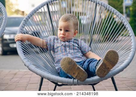 Ginger toddler dressed in jeans and blue t-shirt sitting on open air in wicker chair. Outdoor