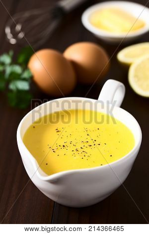 Hollandaise sauce a basic sauce of the French cuisine served in a sauce boat with ground black pepper on top ingredients (egg butter lemon) and whisk in the back photographed on dark wood with natural light (Selective Focus Focus one third into the image)