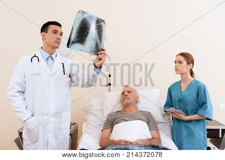 The old man lies on a cot in the medical ward. Next to him is a doctor and a nurse. The doctor looks at the X-ray of the old man. The nurse and the old man also examine the picture.