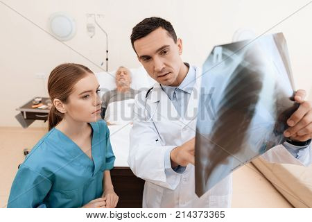 The old man lies on a cot in the medical ward, and next to him there is a doctor and a nurse. The doctor is holding an X-ray in his hands, the nurse is standing next to him. They are in modern clinic.