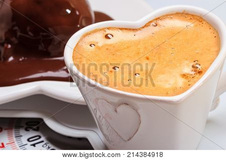 Cup of coffee and chocolate cake on heart shaped plate saucer on weighing scale. Unhealthy nutrition. Fat diet concept.