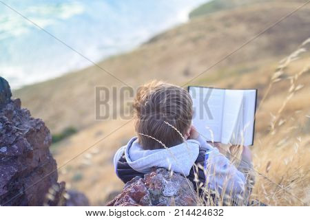 Boy reading book outside on the top of the hill, up above the ocean, unschooling education