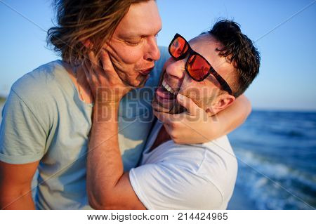 Two men embrace against the background of the sea. They represent a playful kiss.