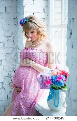 Pregnant woman smiling, looking at belly, holding flowers. Pink dress
