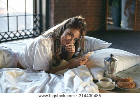 Indoor shot of attractive adult female of Caucasian appearance relaxing on linen on white bed and messaging boyfriend online using home wifi on her mobile phone looking and smiling at camera