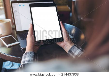 Mockup image of hands holding black tablet pc with white blank screen laptop smartphone and coffee cup on table in cafe