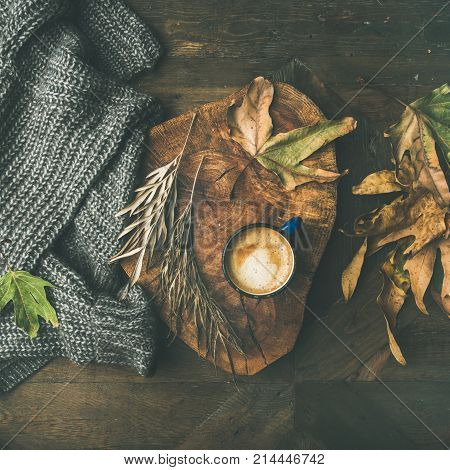 Autumn or Fall morning coffee concept. Flat-lay of knitted woolen grey sweater, wooden tray, mug of coffee and yellow fallen leaves over dark rustic wooden table background, top view, square crop