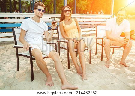 Lifestyle Young people enjoying summer vacation sunbathing drinking at beach bar. Two guys and a young girl sit on the terrace of the summer bar and enjoy the conversation Sun flare