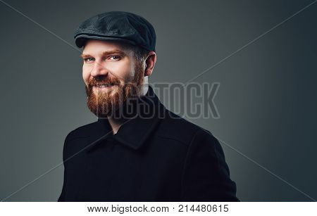 Portrait of Redbeard elegant male dressed in a tweed flat cap and a black jacket over grey background.