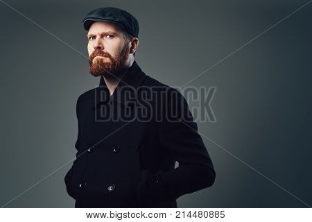Portrait of Redbeard elegant male dressed in a tweed flat cap and a black jacket over grey background.