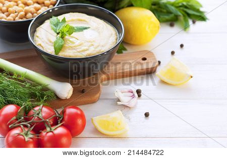 Bowl with hummus, chickpeas, lemon, cherry tomatoes and herbs on a white rustic wooden background.
Middle Eastern cuisine. Background hummus with place for text.