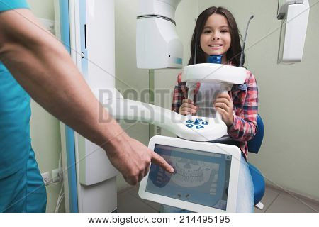 A male doctor makes an x-ray of the girl's jaw, which sits in a special X-ray machine. The girl is smiling. The doctor stands next to him and takes the indicators.