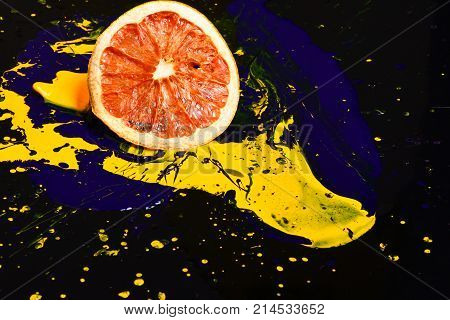 Orange or grapefruit half covered with paints. Paint splashing on grapefruit fruit. Nutrition and food art concept. Drops of blue and yellow oil or acrylic paint poured on fruit on black background.