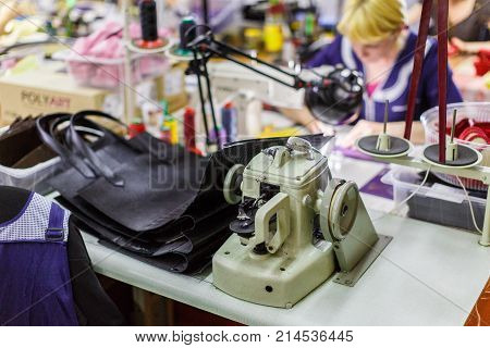 sewing machine and unfinished bags are in focus. blonde woman in dark-blue uniform manufacturing handbags is on the bachground.