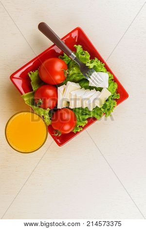 Tomatoes and cheese slices lie on a sheet of fresh salad in a red plate. A glass of orange juice stands side by side on the table.
