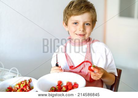 Little blond kid boy helping and making strawberry jam in summer. Funny child cleaning berries and preparing for cooking jam. Kid eating ripe strawberries in domestic kitchen.