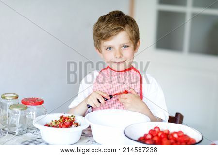 Little blond kid boy helping and making strawberry jam in summer. Funny child cleaning berries and preparing for cooking jam. Kid eating ripe strawberries in domestic kitchen.