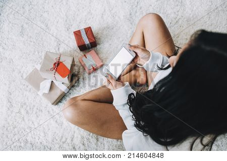 Another view of a girl sitting on the white furry carpet and holding a phone in his hands. She wants to take a picture of Christmas presents that are lying on the carpet. All of them are small but pretty. Close up. Cut view