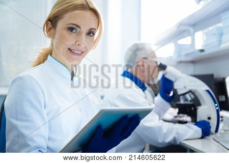 Woman scientist. Good-looking alert smiling budding woman scientist wearing a uniform and holding a tablet while being in the lab and a researcher working with a microscope in the background