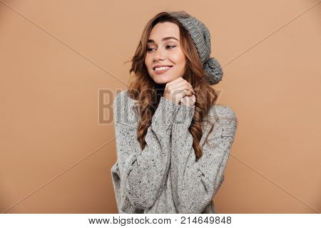 Charming young brunette woman in warm clothes holding hands, looking aside, isolated on beige background