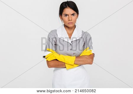 Portrait of grumpy attractve maid in uniform and yellow rubber gloves standing with crossed hands and looking at camera, isolated on white background
