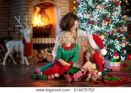 Family with kids at Christmas tree and fireplace. Mother and children opening gifts at fire place. Boy girl and mom open presents. Winter holidays interior decoration. Child in pajamas on Xmas eve.