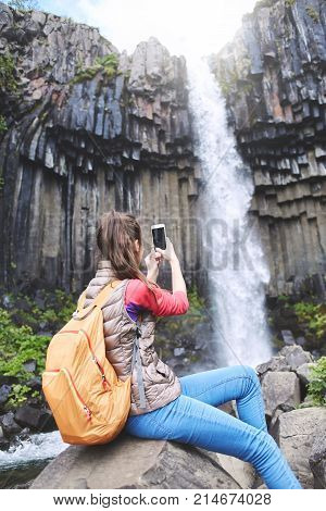 Woman tourist photographing on smartphone Svartifoss waterfall surrounded by basalt columns in the south of Iceland. Skaftafell national park in Iceland