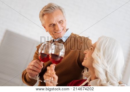 Love you to the moon and back. Selective focus on a happy retired man smiling to his wife while both enjoying their glasses with red wine and celebrating anniversary together.