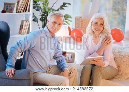 Recharging batteries. Positive minded senior couple looking into the camera with cheerful smiles on their faces while relaxing at home with a glass of wine and watching videos on a touchpad.