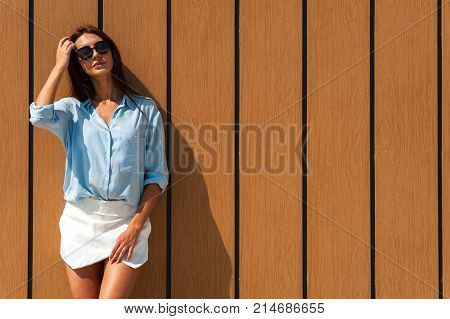 Young beautiful tanned brown-haired woman in sunglasses posing against wooden fence in the sun on warm summer day