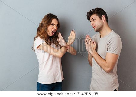 Portrait of a young casual couple standing together isolated over gray wall background, man asking for something and woman refuses
