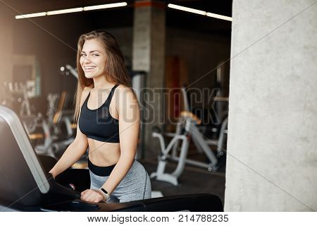 Young female body fitness coach running on a threadmill looking at camera smiling using monitor to track heart rate. Athletic trainer warming up early in an empty gym.