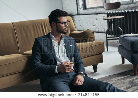 Enjoying favorite drink. Handsome young man in full suit holding a drinking glass and looking away while sitting on the floor at home