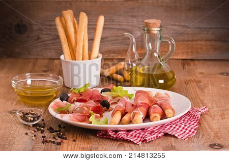 Grissini breadsticks with ham and olives on white dish.